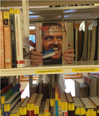 Bok med ansiktet til skuespiller Jack Nicholson fra filmen Ondskapens hotell, holdes av hender inni en bokhylle.