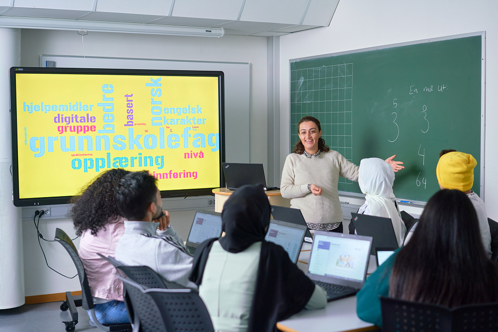 Et klasserom med en lærer som peker på en tavle med matematiske utregninger. Foran læreren står en stor skjerm med et ordskyer med ord som "grunnskolefag", "opplæring", "norsk", "engelsk", "digitale hjelpemidler" og "nivå". Flere elever sitter med bærbare datamaskiner og ser mot skjermen eller læreren.