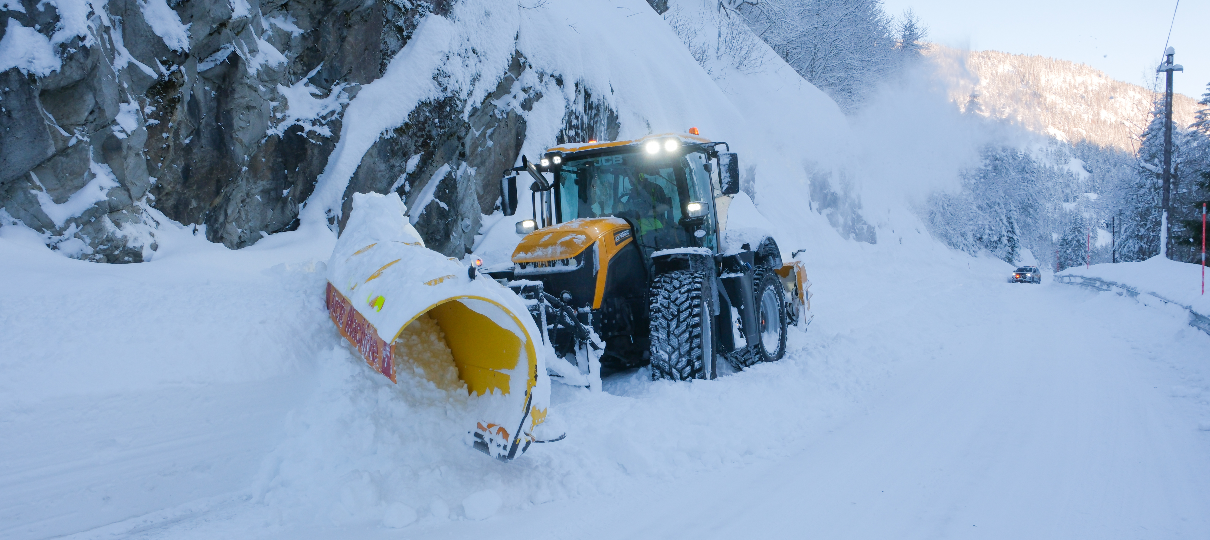 Hjullaster med brøyteplog og snøfres i arbeid mot kamera. Fjell skimtes i bakgrunnen.