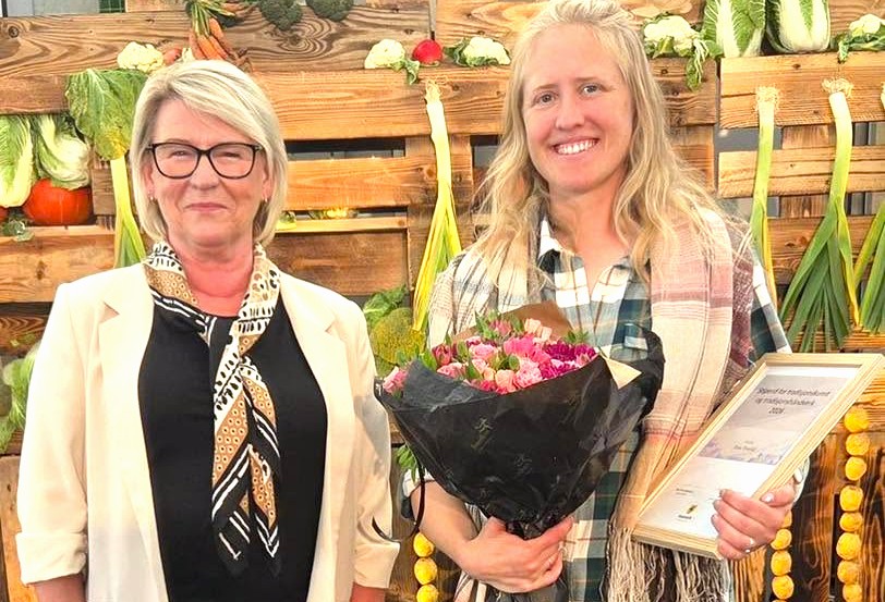 To damer smiler til kamera, en av dem holder en blomst og et diplom.