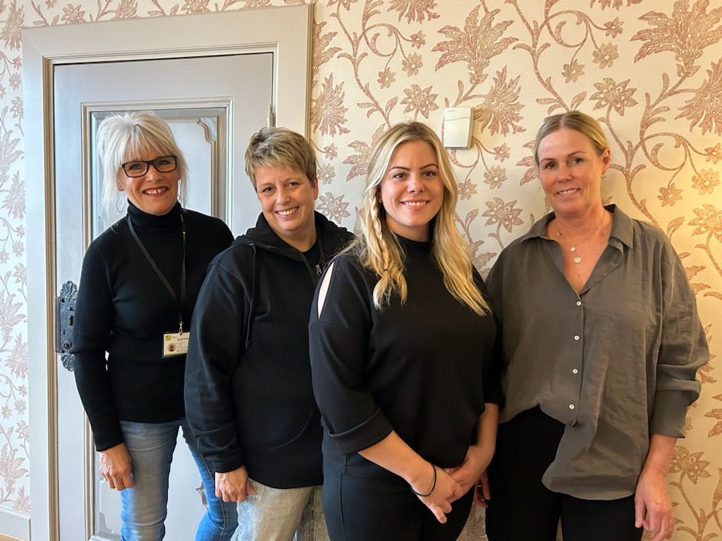 Fire kvinner står foran en vegg med blomstrede tapet.