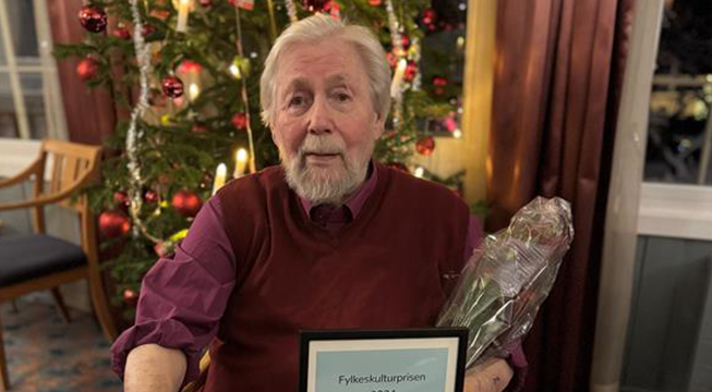 Mann i rød skjorte og strikkevest som sitter med diplom og en blomst.