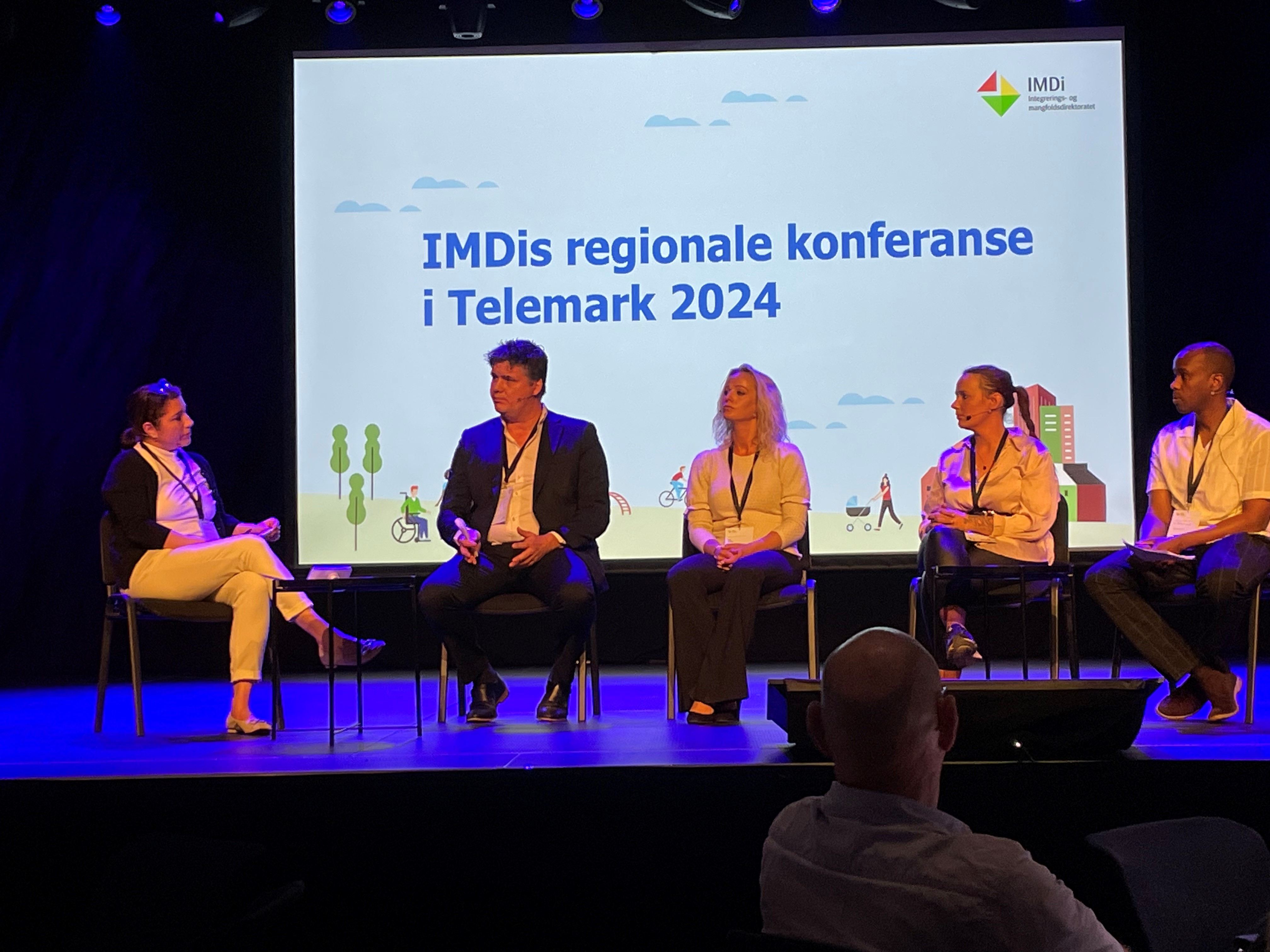 Fem personer på en scene med en skjerm bak hvor det står "IMDis regionale konferanse i Telemark 2024"