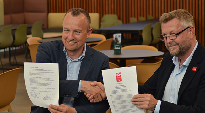 To menn som smiler og tar hverandre i hånda mens de holder opp to hvite ark med tekst