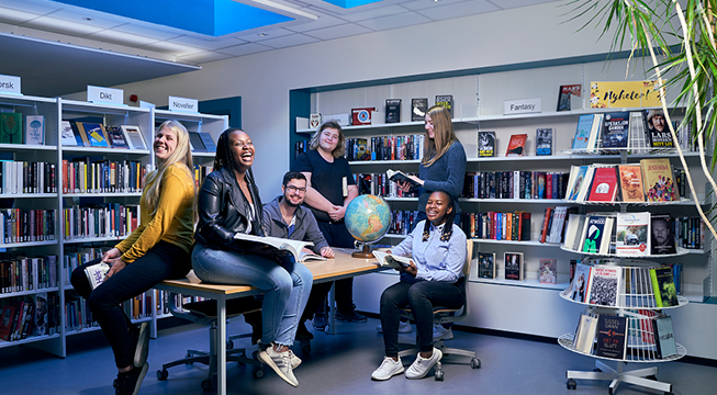 Seks elever sitter i skolens bibliotek med bøker i hendene og smiler mot kamera