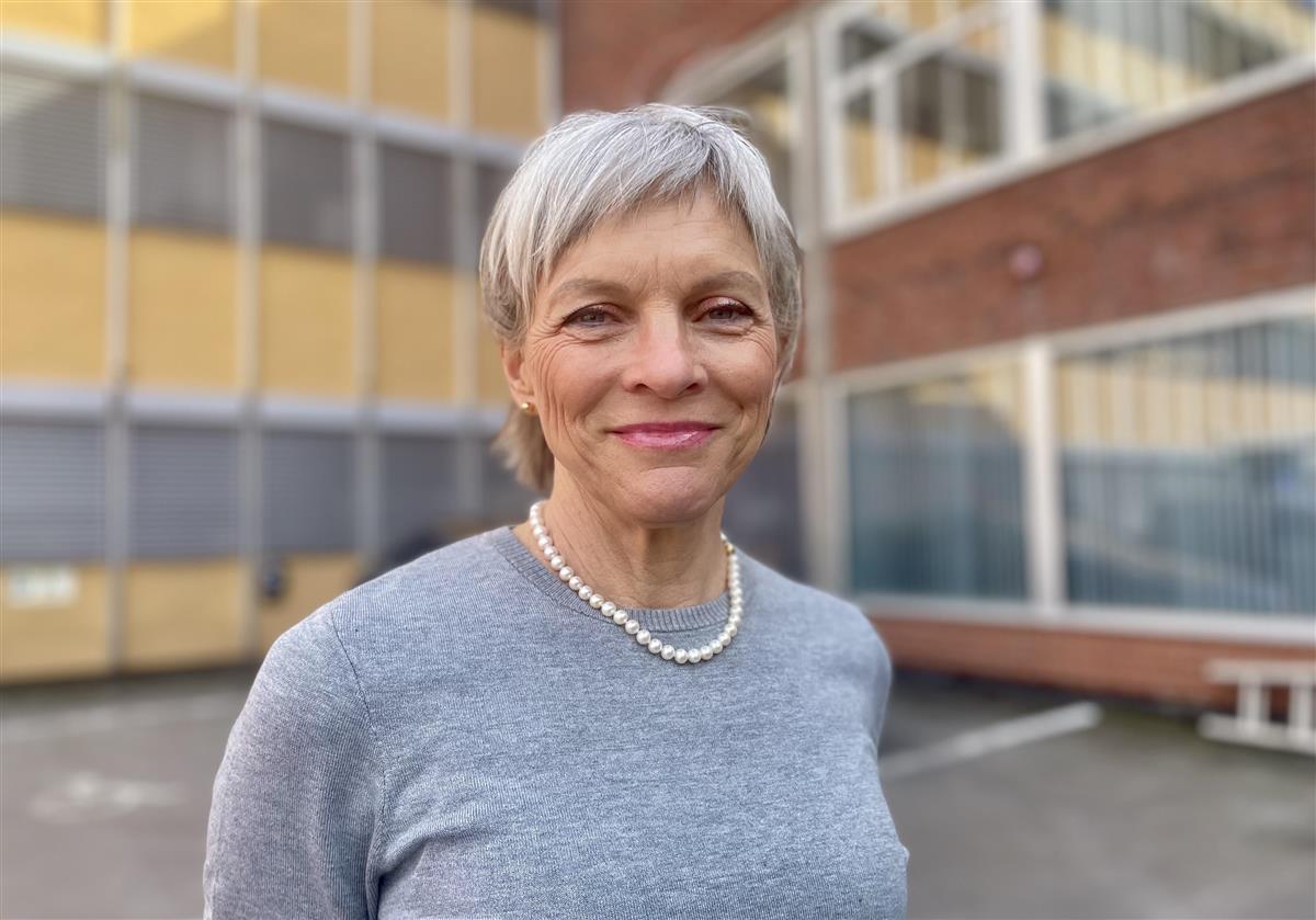 En smilende kvinne med kort, grått hår og grå genser. Et stort bygg i bakgrunn.