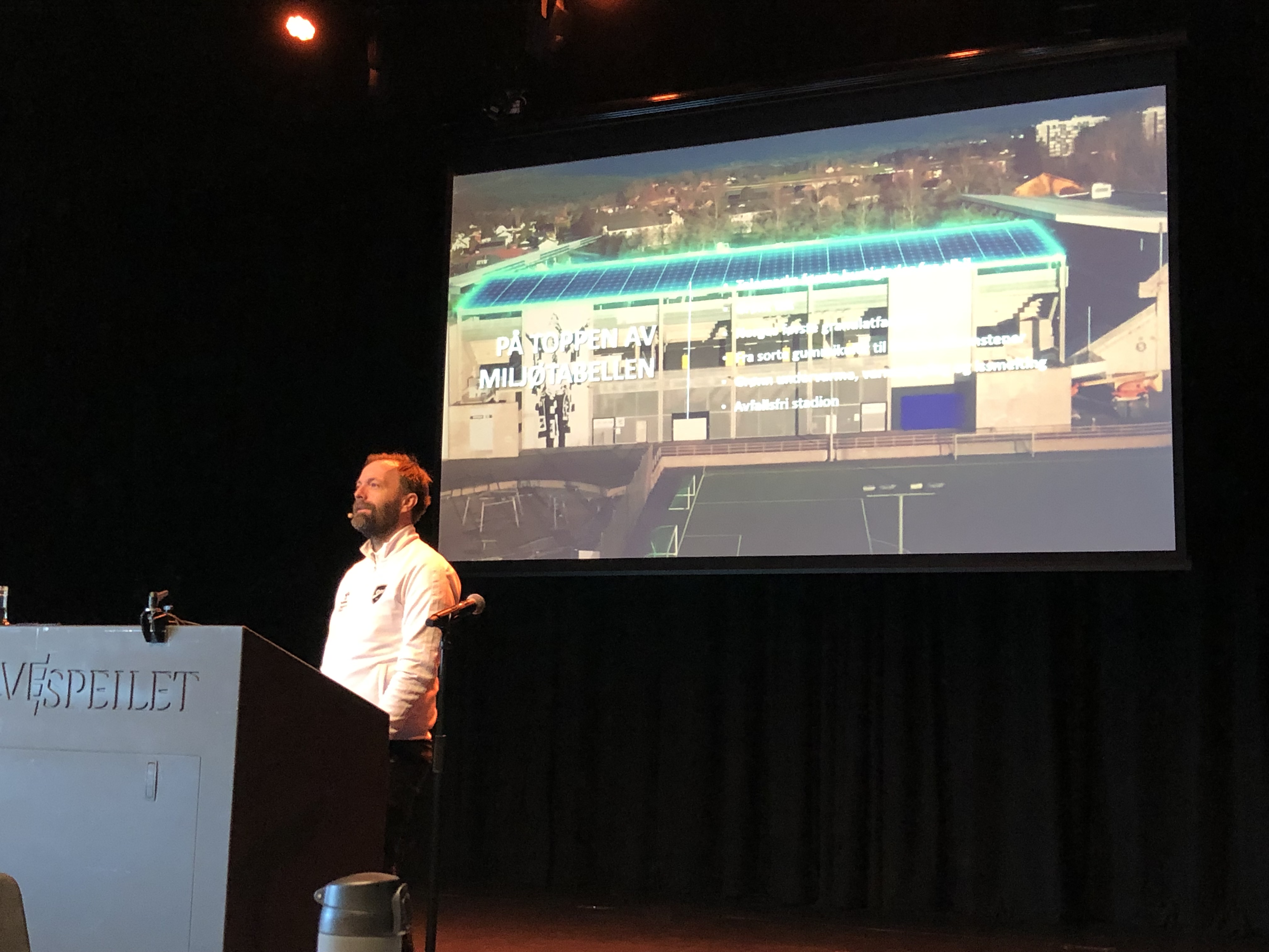Mann på scenen i hvit jakke foran en skjerm.