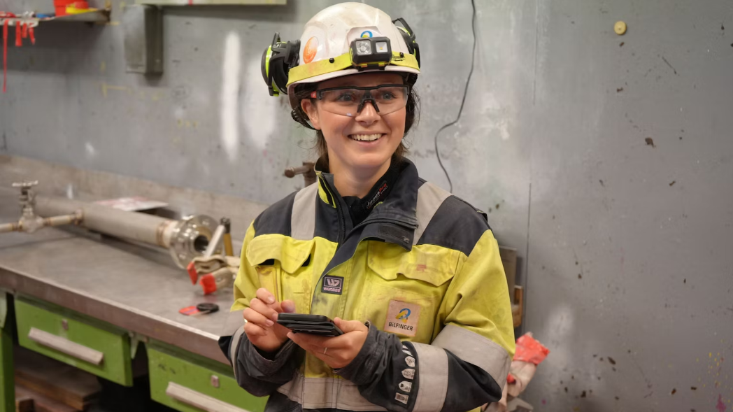 Kvinne med arbeidsklær i sort og gult, hjelm og briller som smiler.