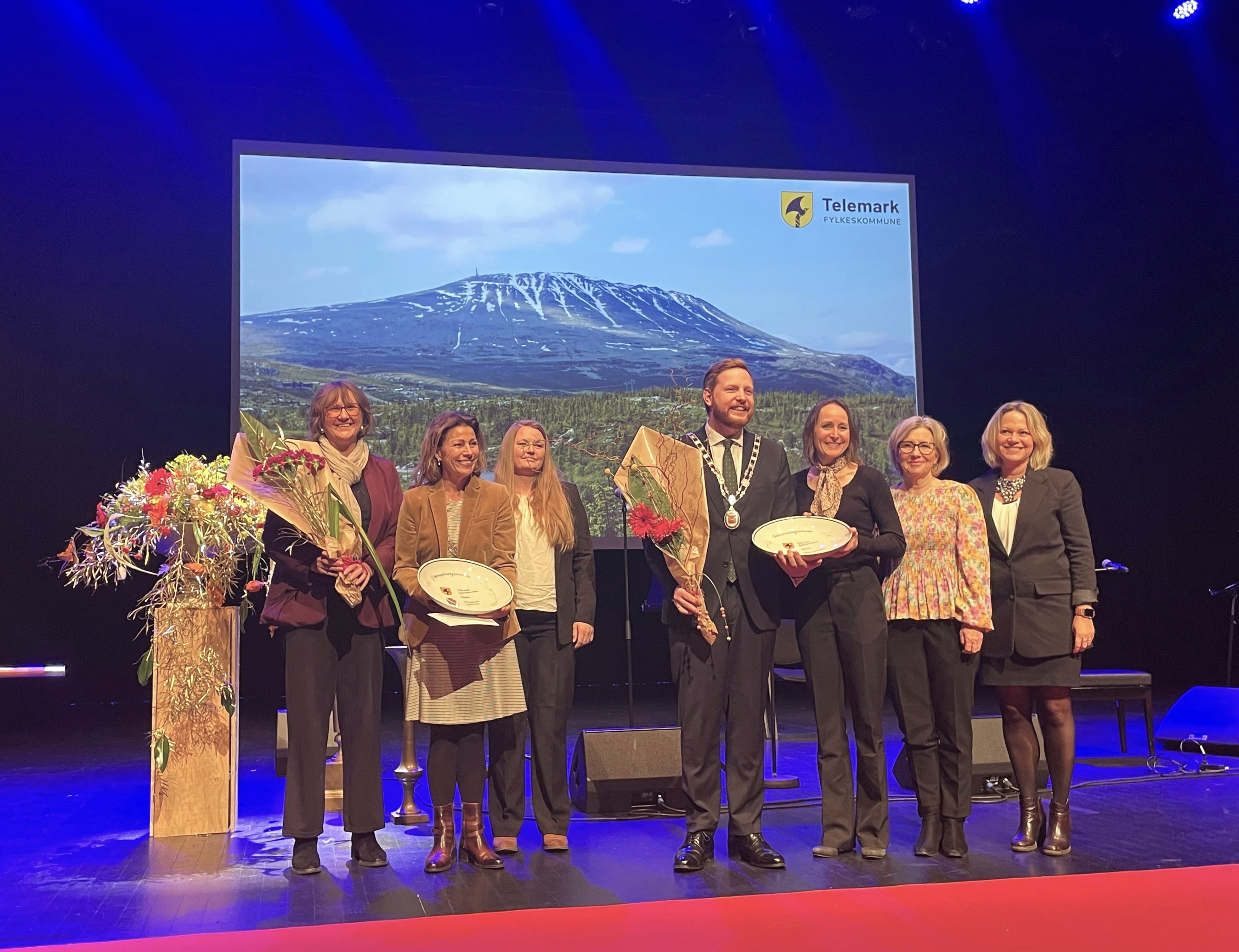 7 personer på en scene. De er pent kledd. To av dem holder blomster og to av dem holder store, runde plaketter. Alle personene smiler.